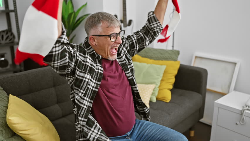 Joyful middle-aged man with glasses cheering with a sport scarf in a cozy living room.