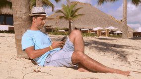 Young attractive man sitting under a palm tree on beach by sea on summer vacation, holding using internet on smartphone, dressed in hat. Young man tourist sitting on a beach sand with a smartphone - Powered by Shutterstock - Get 15% off with code: PIKWIZARD15