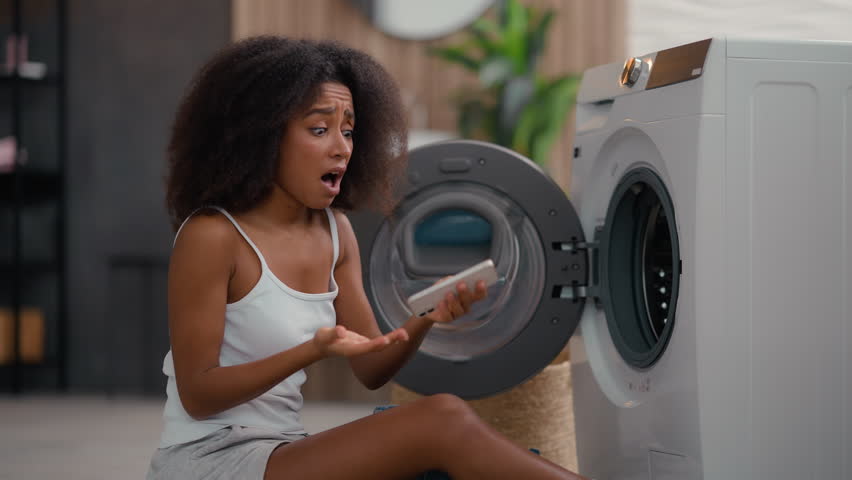 African American girl ethnic lady biracial woman housewife home laundry clothes open washing machine housekeeping take clean wet jeans pants find mobile phone in pocket broken smartphone shock upset