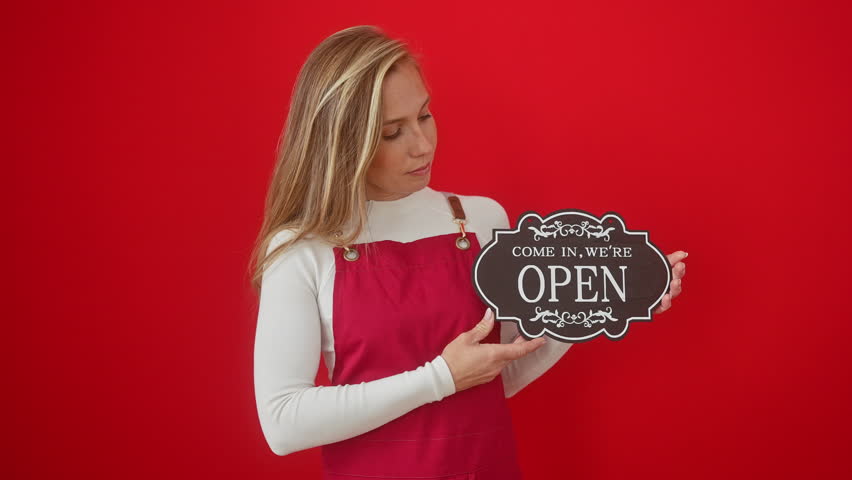 Joyful young woman, confidently smiling with teeth, blonde in apron, holding blackboard, over red isolated background