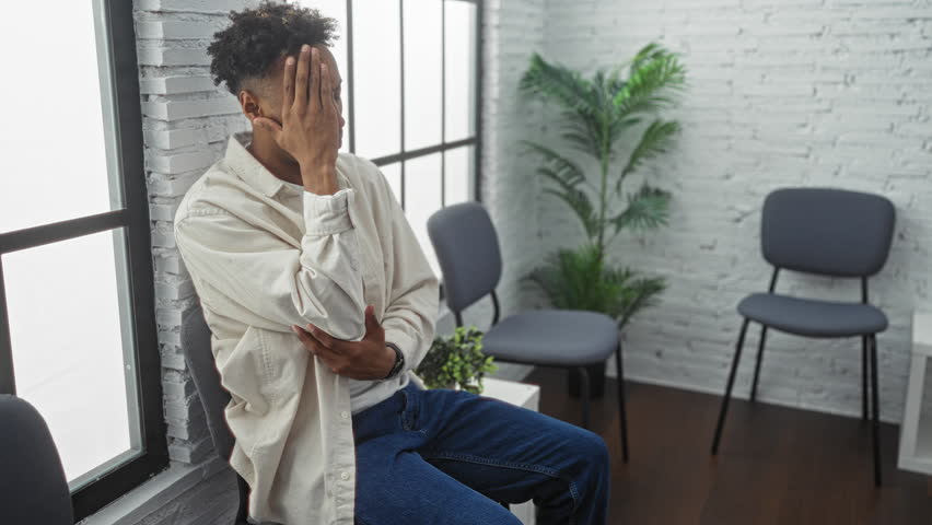 Handsome young african-american man with a beard sitting in an indoor waiting room with plants and empty chairs, exhibiting a thoughtful expression in a modern, minimalist interior.