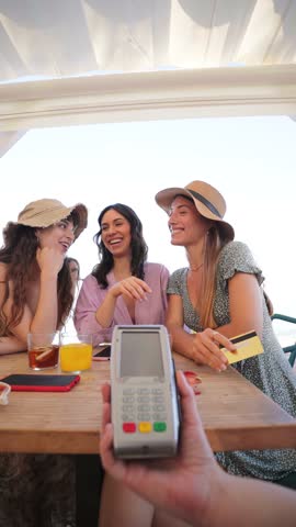 Vertical. Happy young group of only women paying the bill with a contactless credit card in cocktail beach bar. Female smiling holding a creditcard and giving a payment transaction to the cashier
