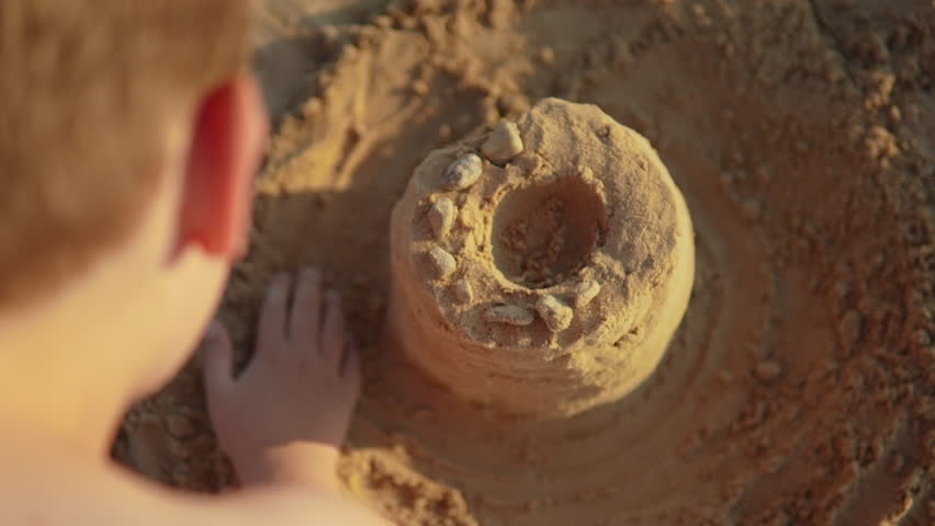 Child builds sand castle tower on beach, viewed over the shoulder. Boy strengthens fantasy building with sand at sunset. Childhood and carefree summer holidays.