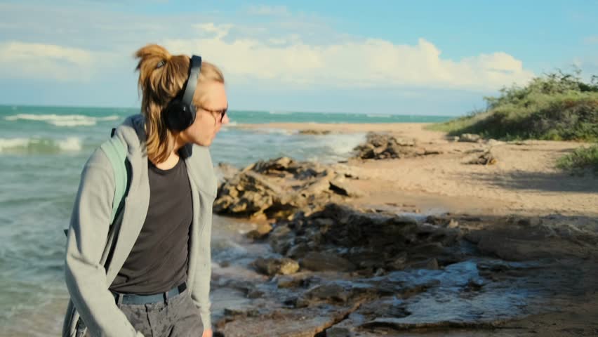 Romantic student dancing on the beach with a backpack and headphones, squinting from sun against the backdrop of a picturesque seashore.
