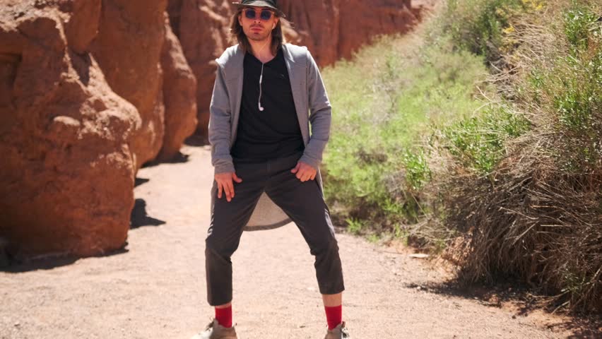 Young man dancing near sandy stone in hipster clothes swaying to the rhythm of music on summer trip, performs the snake dance.
