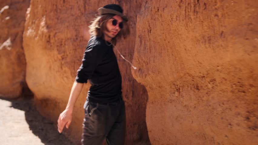 Extravagant dance of a young guy in a black hipster attire circling against the backdrop of a sandy mountain slope, slow motion.