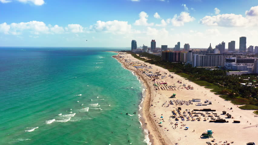 4k Gorgeous aerial footage. Perspective of Miami Beach reveals its turquoise waters, sandy shores with sunbathers, and the city skyline, offering a tranquil vacation experience by the Atlantic Ocean