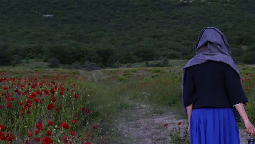A woman walks along the road along a poppy field at dusk with a kerosene lamp in her hands against the backdrop of the mountains