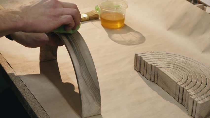 A hand applies oil to a curved wooden piece using a sponge. Other parts of the same shape are laid out on the table. Nearby are a brush and a container of oil, prepared for wood treatment