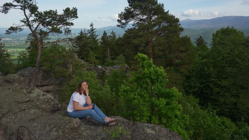 Young woman in a white T-shirt sitting on a peak and relaxing. Bird watching against the background of a beautiful mountain landscape. Relaxing and resting after a hike through the mountains. Hiking. 