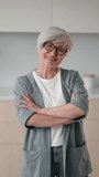 Happy grandmother with eyeglasses looking at camera joyfully, vertical video, portrait. Beautiful senior woman with grey hair and wrinkled face folded hands on chest, positive emotions and good mood