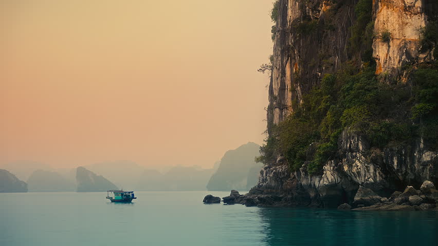 A breathtaking nature view of a boat on the sea near a rocky cliff at Ha Long bay under foggy orange sunset sky in Vietnam