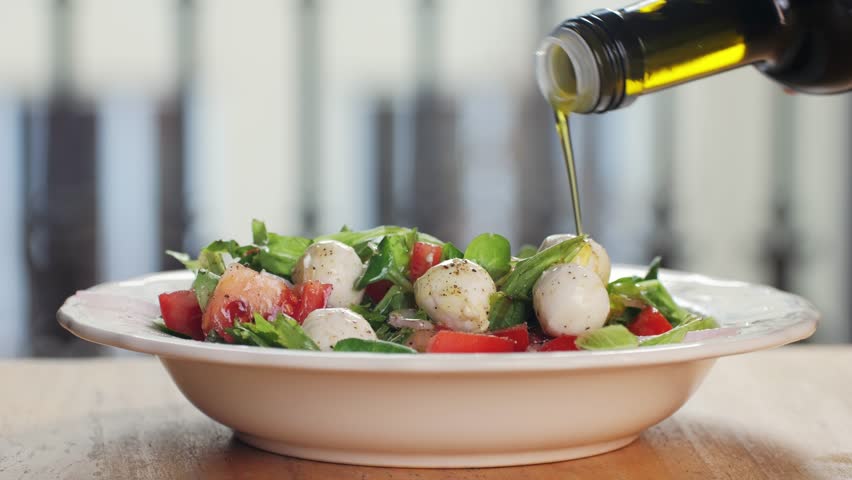 Pouring olive oil in Fresh Caprese salad on white plate close-up. Caprese is an Italian salad, made of sliced fresh mozzarella, tomatoes, and sweet basil, seasoned with salt, and olive oil. Food