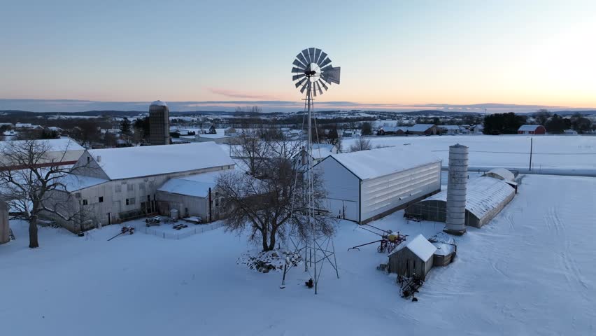 Dawn lights up a snow-covered farm with barns, a silo, and a classic windmill, embodying a serene rural winter scene. Aerial orbit during sunrise.