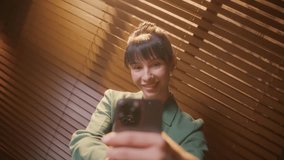 Portrait of beautiful brunette woman laughing and showing peace sign while taking selfie photo on cellphone on background roller blinds. Businesswoman taking selfie with mobile phone - Powered by Shutterstock - Get 15% off with code: PIKWIZARD15