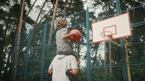 Rear of Caucasian family dad and kid boy having fun playing basketball. Happy son sitting on father's shoulders throwing a ball in hoop outdoors on court. Leisure activity. Bottom view. Sport concept - Powered by Shutterstock - Get 15% off with code: PIKWIZARD15