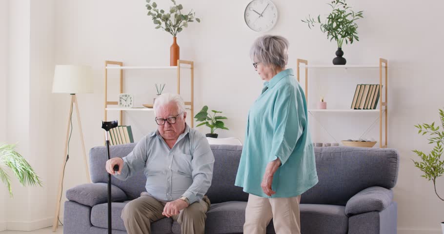 Married senior couple overcoming health problems together. Senior woman helps sick weak man to stand up from sofa and walk slowly with crutch in living room. Disability, care, assistance, support