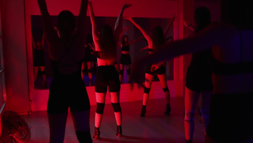 A group of girls in shorts stand in front of a mirror, doing exercises and stretching their bodies. Dance class in red lighting. Group dance.