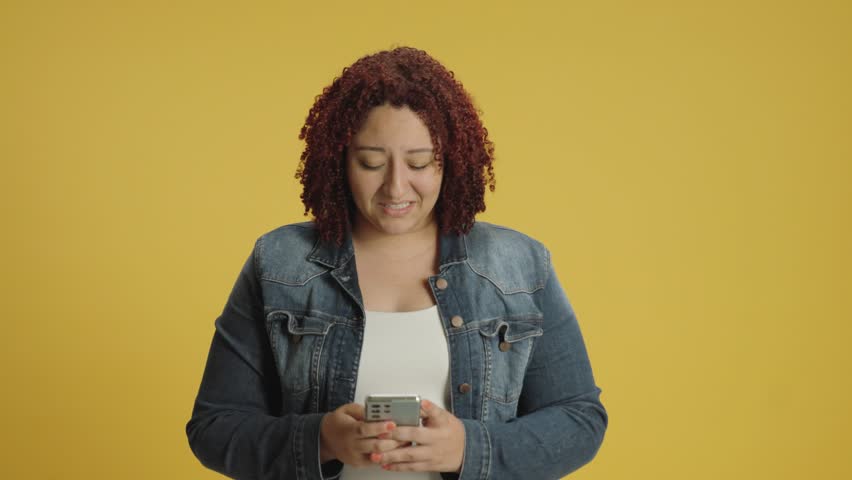 Happy young hispanic woman 20s wear denin jacket and white shirt hold use mobile cell phone typing say wow yes just found out great big win news doing winner gesture isolated on yellow background stud