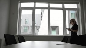 A woman in a conference room, checking her phone and pacing by the window, embodies patience and professionalism in a business environment. - Powered by Shutterstock - Get 15% off with code: PIKWIZARD15