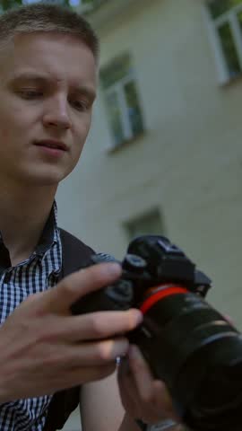 Young guy tourist sits in an alley on a bench and scrolls at the photographs taken on a digital expensive big photo camera. Casual clothes with a black backpack. Low angle close-up. Vertical shot.