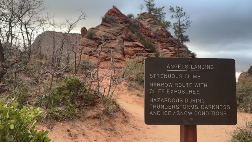 A warning sign marks the trailhead for the challenging Angels Landing Hike in Zion National Park, Utah. The iconic red rock formations and desert landscape of Zion Canyon loom in the background.