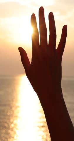 Female hand touching the sun, sunlight passing through fingers at sunset on the sea, slow motion closeup shining sun and the sea vacation, season travel nature idyllic