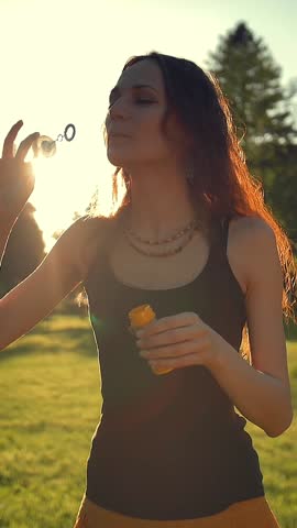 Portrait of beautiful redhair young woman. Blowing bubble in the park. Super slow motion shot in 180 fps. Sunrise or sunset straight orange light. Vertical shot.
