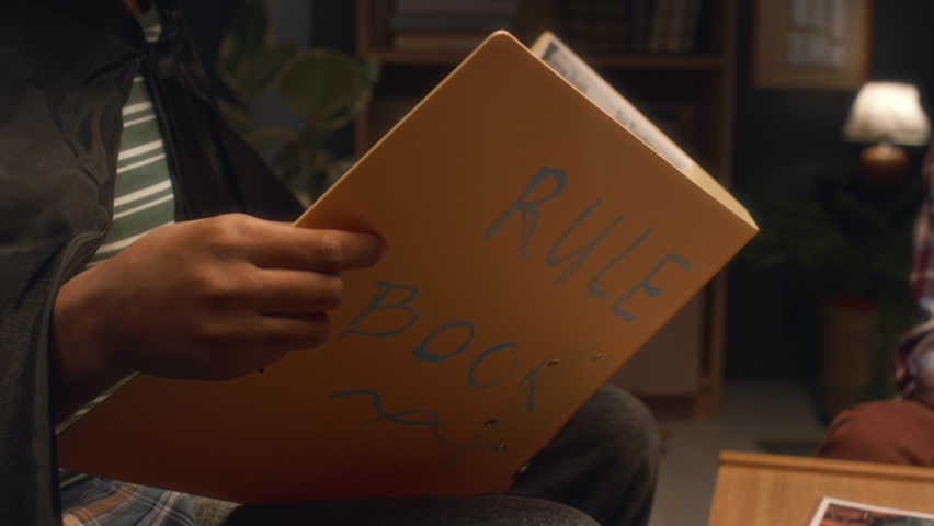 Side tilt shot of Black boy as game master dressed in wizards costume and holding large folder with Rule Book inscription while role playing with friends at home in evening