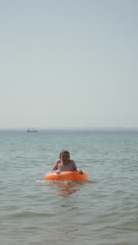 Joyful Caucasian boy is having fun jumping with splashes with bright inflatable ring in sea with ship. Vertical video of happy child having fun on vacation at resort with personal safety equipment.