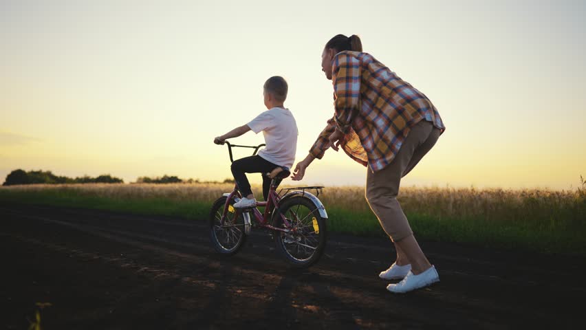 Young woman teaching son to ride bicycle in field in sunset, happy childhood. Joint family time in summer vacation, active pastime in nature, little boy riding bike, mother helping to his child