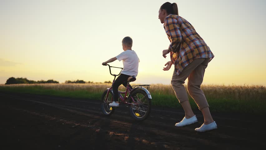 Young woman teaching son to ride bicycle in field in sunset, happy childhood. Joint family time in summer vacation, active pastime in nature, little boy riding bike, mother helping to his child