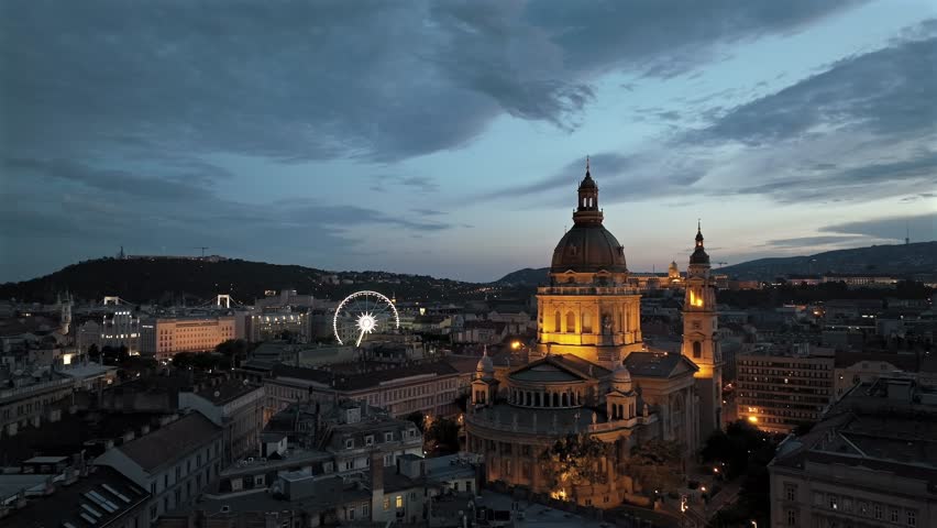 Aerial video about illuminated  St Stephens basilica at dusk. Budapest eye ferris wheel, Buda royal castle and Danube river is on the background
