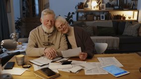 Medium zoom-in portrait of happy loving senior Caucasian husband and wife sitting together in cozy living room, head on shoulder, smiling, while managing financial affairs, paying taxes and bills - Powered by Shutterstock - Get 15% off with code: PIKWIZARD15