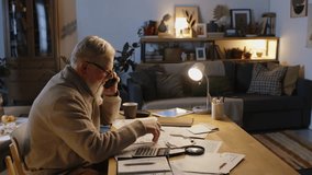 Medium pan shot of 70-year-old Caucasian male pensioner sitting at desk in living room, working with bills, financial documents, talking on phone with electricity provider to dispute wrong charge - Powered by Shutterstock - Get 15% off with code: PIKWIZARD15