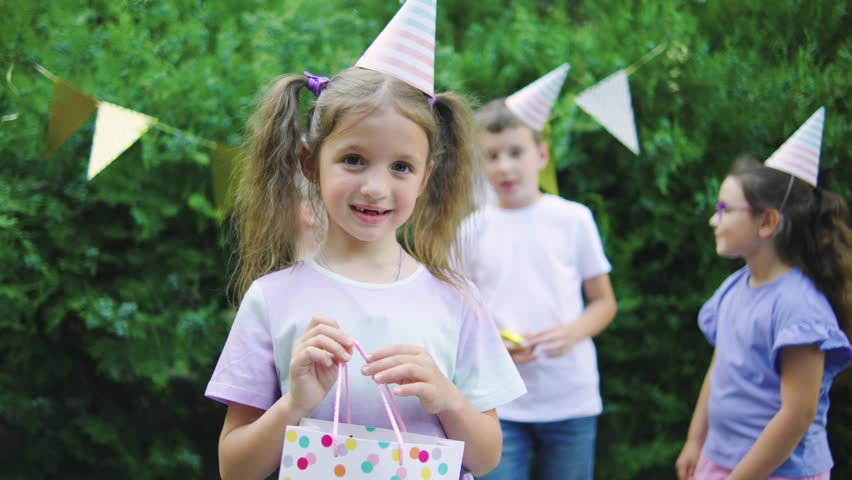 Birthday girl in colorful t-shirt holding her birthday gift and smiling, outdoors birthday party with happy kids and holiday decorations in the background