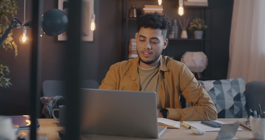 Cheerful Arab man talking and waving hand during online video call using laptop in apartment in evening. Modern technology and communication concept.