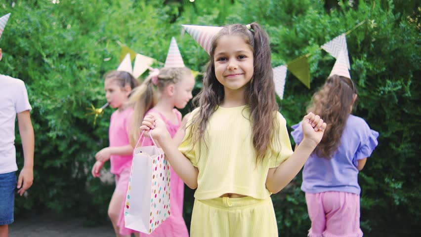 Birthday girl in colorful t-shirt holding her birthday gift and smiling, outdoors birthday party with happy kids and holiday decorations in the background