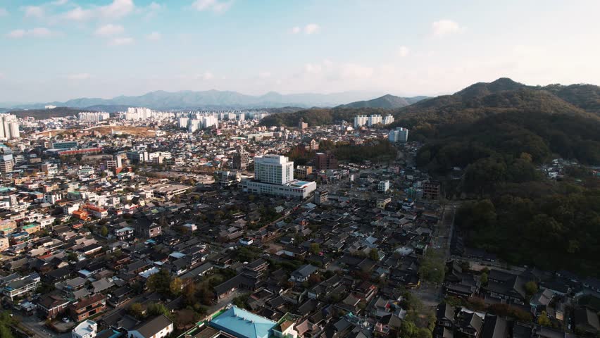  Aerial view of Jeonju Hanok Village showcases traditional Korean houses with tiled roofs, juxtaposed against modern buildings, encircled by verdant hills, reflecting a harmonious cultural landscape.