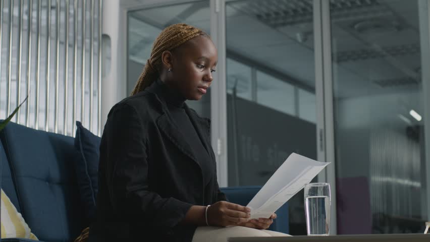 Young woman in businesswear drinking water and reading while waiting for colleague in office