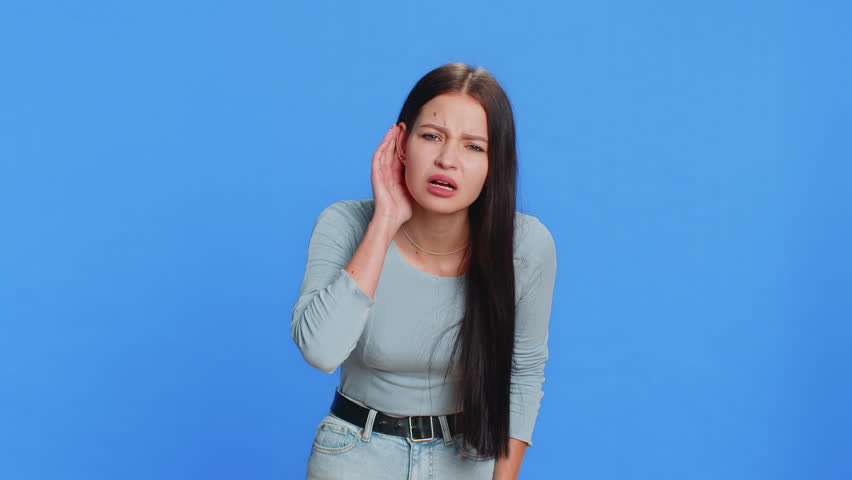 I can't hear you. What. Young woman trying hear you, looking confused and frowning, keeping arm near ear for louder voice, asking to repeat, to hear information, deafness. Pretty brunette adult girl