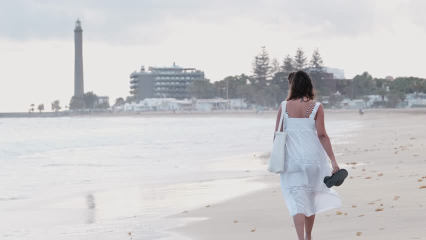Woman walking along the beach at sunset wearing a white dress. Concept: outdoors, enjoy, disconnection
