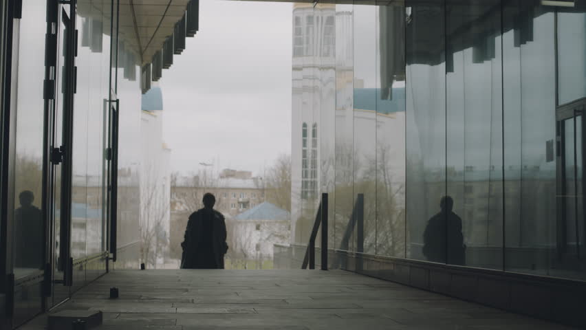 Full shot of young Asian man in long trench coat walking between two glass walls on city street