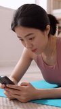 Vertical HD video of smiling young asian woman using mobile phone while relaxing after exercising at home lying on mat. Sport, technology and healthy lifestyle concept.