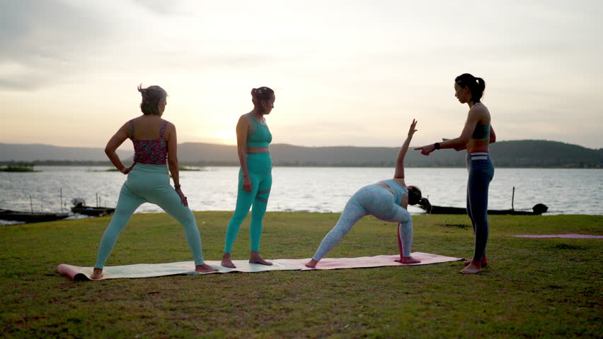 A group of women are practicing yoga on a grassy field by a lake. The sky is cloudy and the water is calm