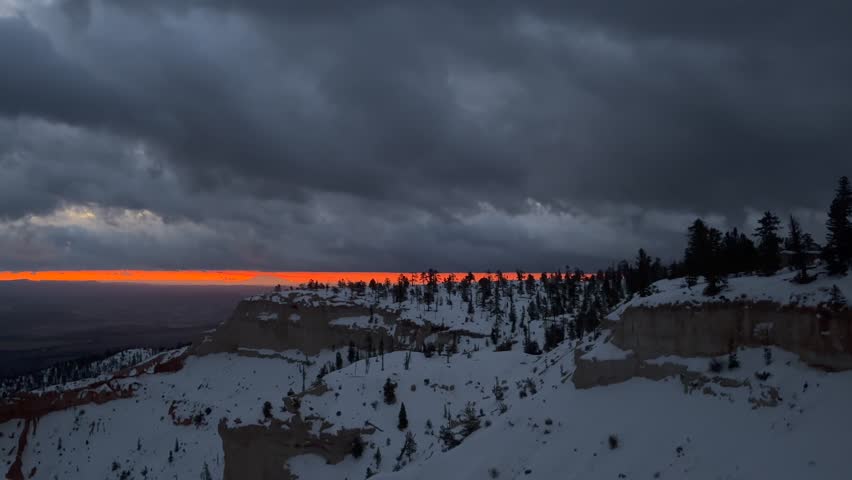 Dawn breaks over the snow-covered landscape of Bryce Canyon National Park in Utah. A fiery orange sunrise paints the horizon beneath dramatic storm clouds, creating contrast of light and shadow - USA