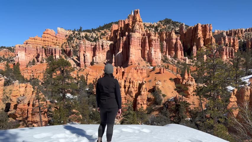 A woman enjoys the winter views of Bryce Canyon National Park in Utah, USA. She is looking up at the red rock hoodoos, pine trees, and snowy landscape from the canyon on a bright sunny day.