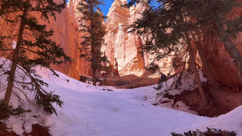 A male hiker trekking through the snow on the Peekaboo switchbacks in Bryce Canyon National Park, Utah. Sunlight hits the top of the red rock hoodoo formations - USA