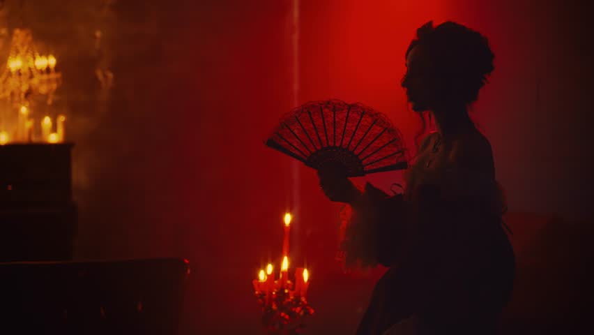 Art portrait dark shadow silhouette gothic sexy fantasy woman vampire queen holding in hands black lace fan in red light candles burning flame. Vintage dress Mystery noble lady girl night club room 4k