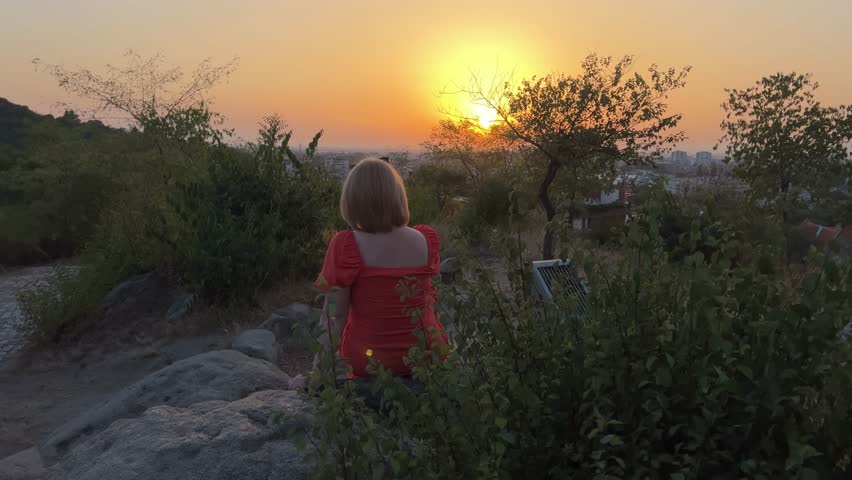Woman sitting on a rock enjoys sunset over the city of Plovdiv on Sahat Tepe. Beautiful orange sunset. Dusk. Golden hour.